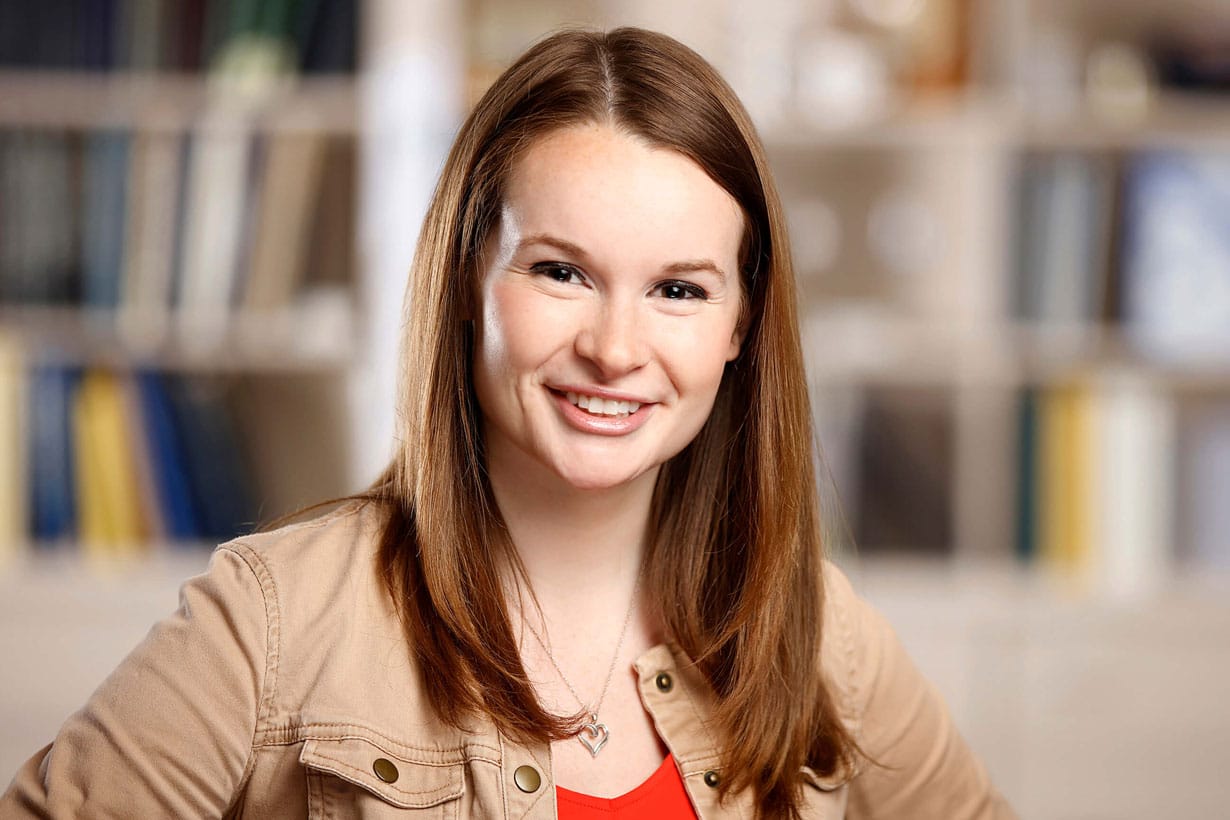 A person with long brown hair, wearing a beige jacket and red top, smiles at the camera.