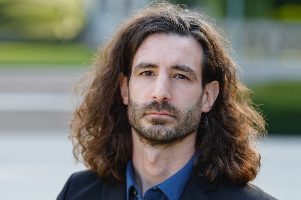Person with long brown hair wearing a dark blazer and blue shirt, outdoors.