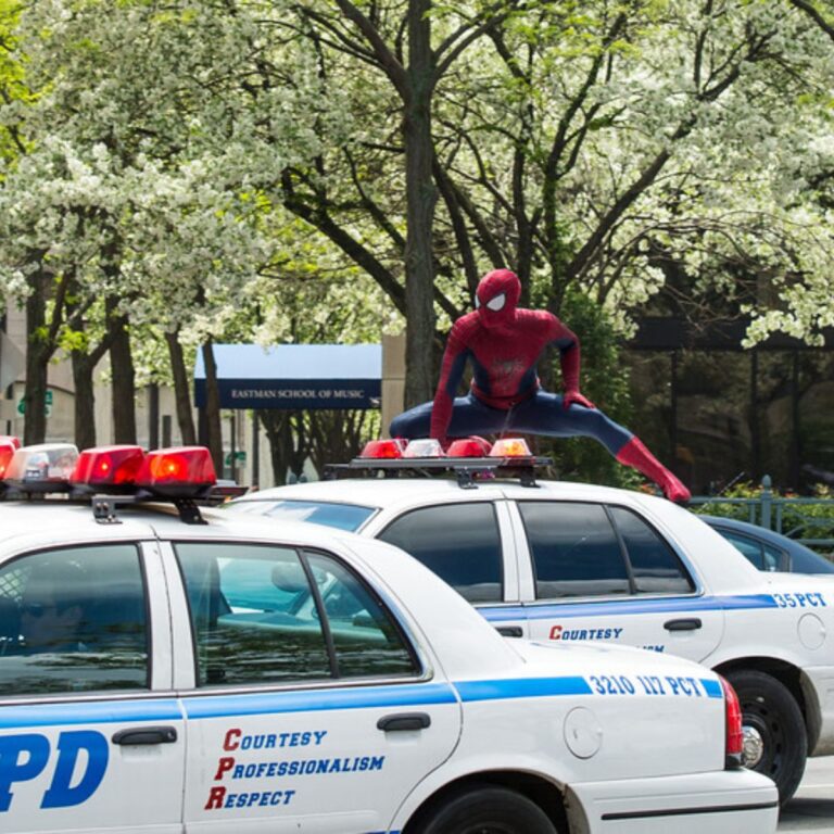 A costumed figure crouches atop a police car with blooming trees and a school sign in the background.