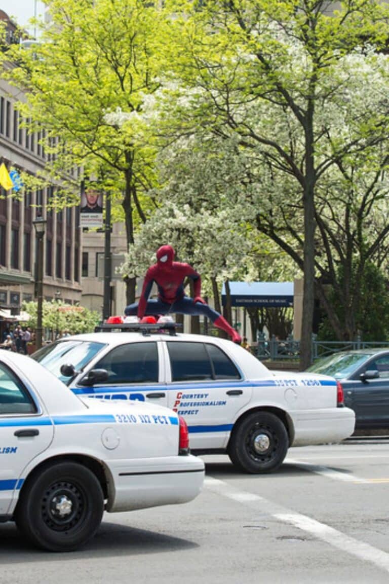 Spider-Man crouches on a police car in a city street with buildings and trees.