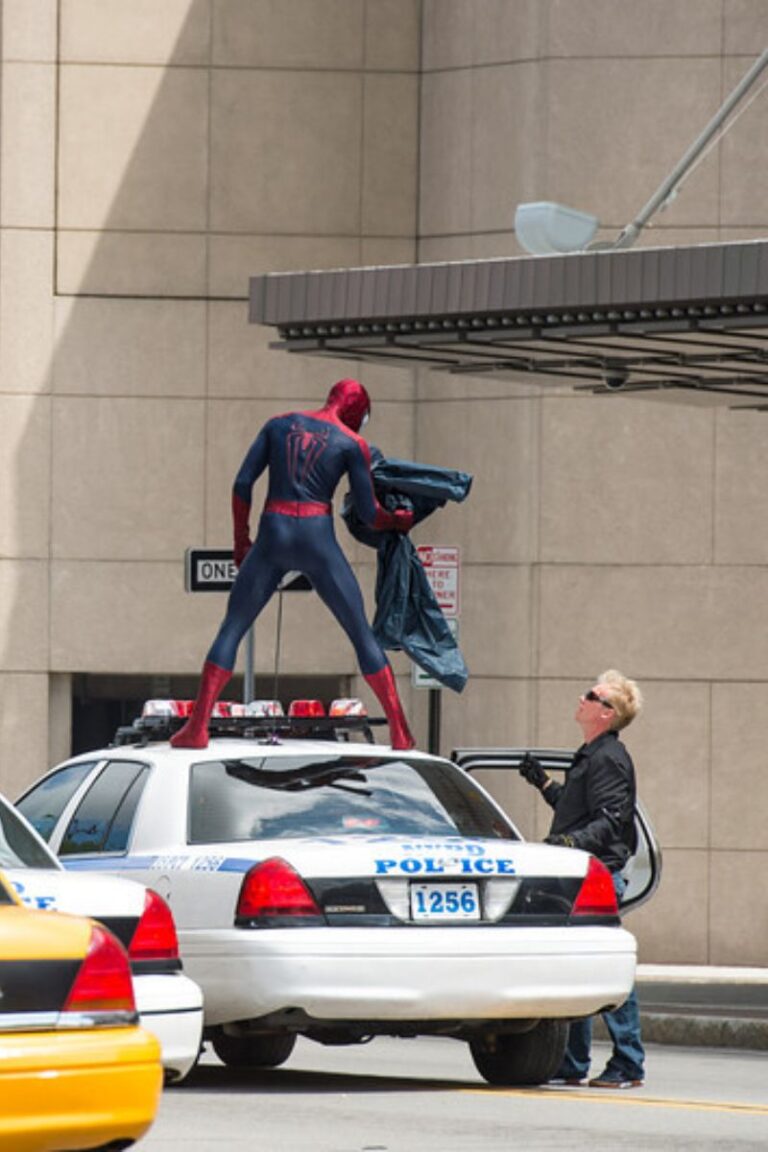 Spider-Man costumed person standing on a police car while another man looks up.