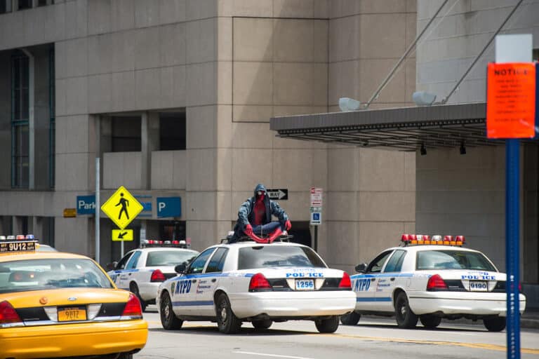 Spider-Man crouched on an NYPD police car roof surrounded by police cars and a yellow taxi on a city street.