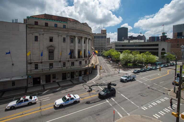 Urban scene with a neoclassical building, police cars, and a camera truck on the street.