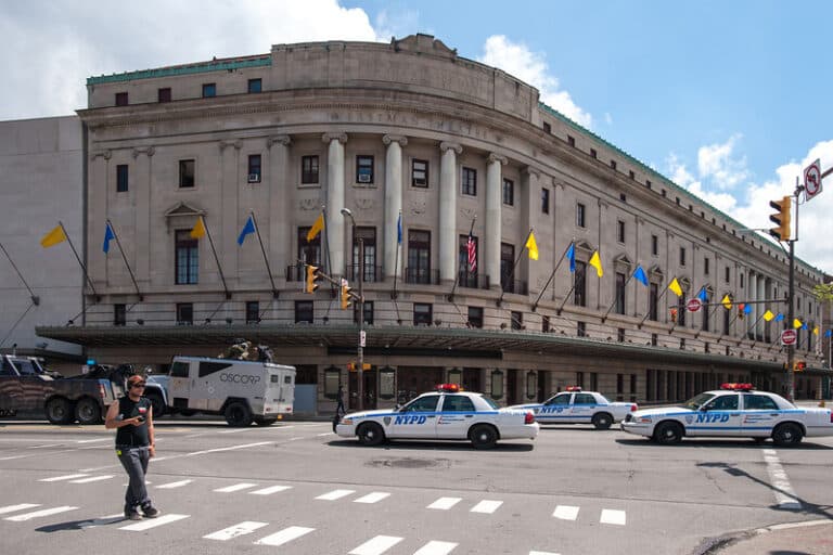 Historic building with NYPD cars and OSCORP truck in front; blue and yellow flags line the structure.