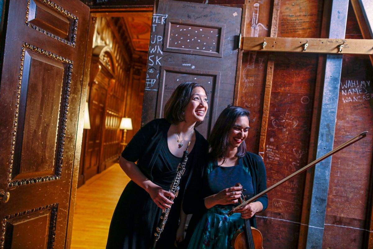 Two women stand in a room at the University of Rochester's Eastman School of Music, one holding a violin near a door.