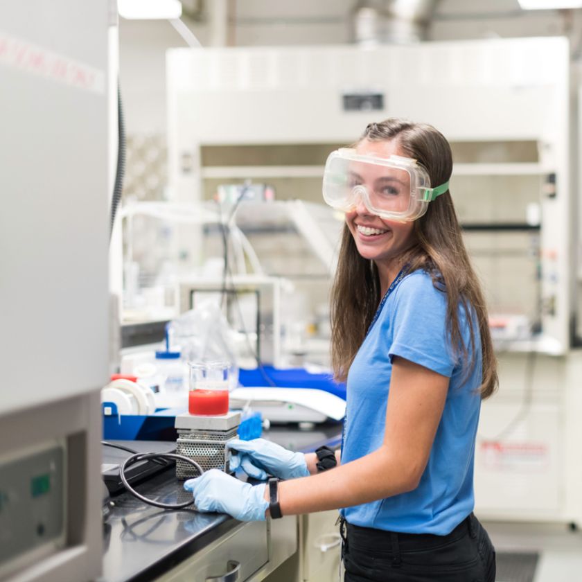 A University of Rochester student in a lab coat and goggles, smiling while working in a laboratory setting.