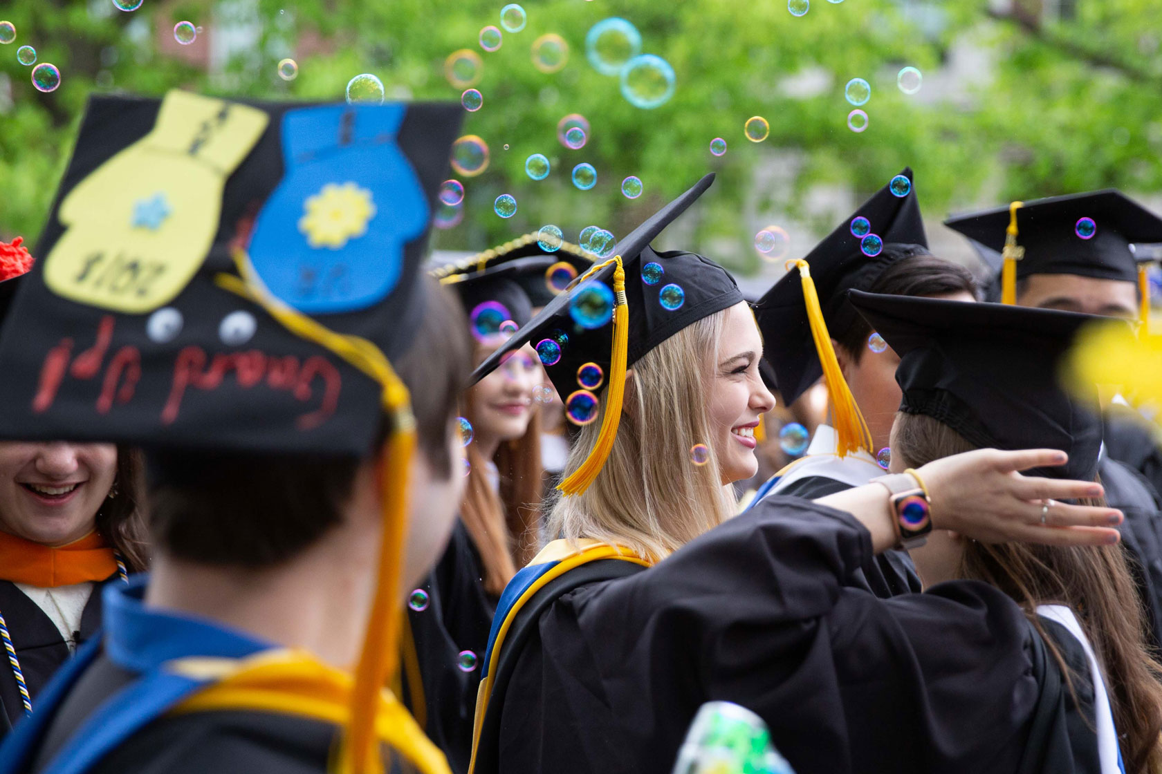 Graduates in caps and gowns celebrate at the University of Rochester, surrounded by floating bubbles in the air.