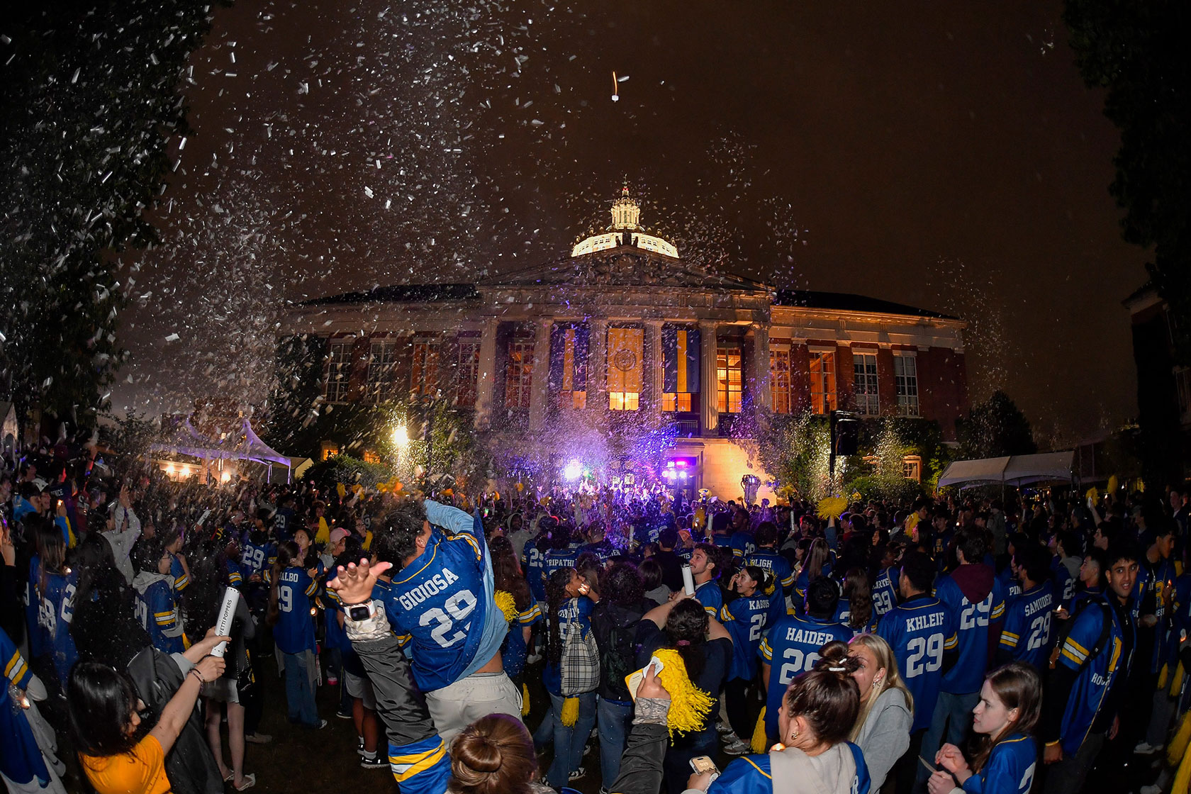A crowd of people celebrating with confetti in front of the University of Rochester building.