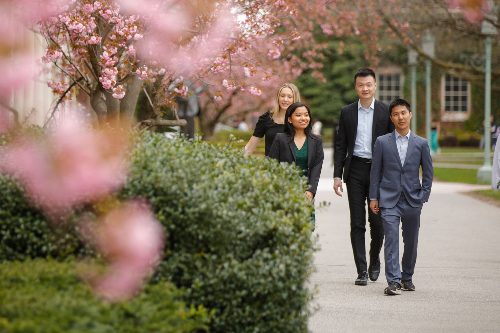 Three people stroll along a sidewalk adorned with pink flowers at the University of Rochester.
