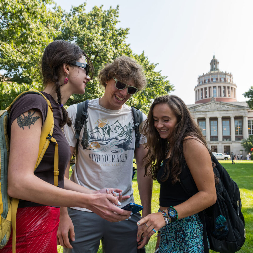 Three students looking at a smartphone in front of a large building with columns and a rotunda.