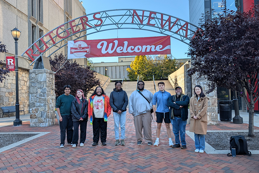 A group of eight people pose in front of metal arch and buildings.