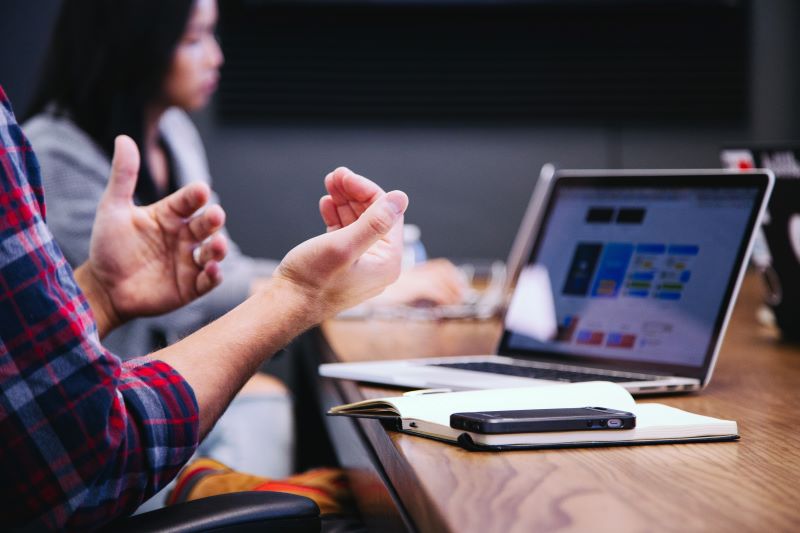 Person using hand gestures during a conversation while working on a laptop with another person in the background.