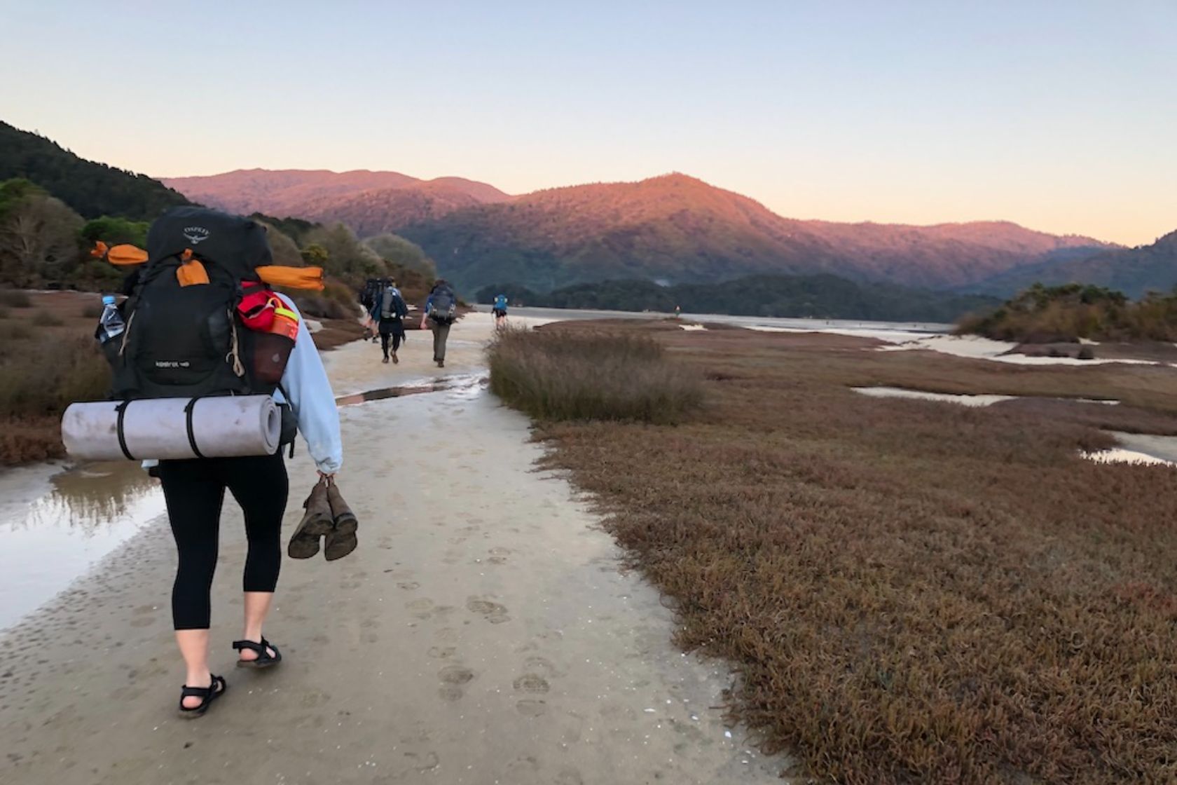 Hikers walk along a sandy path through wetlands towards distant mountains at sunrise or sunset.