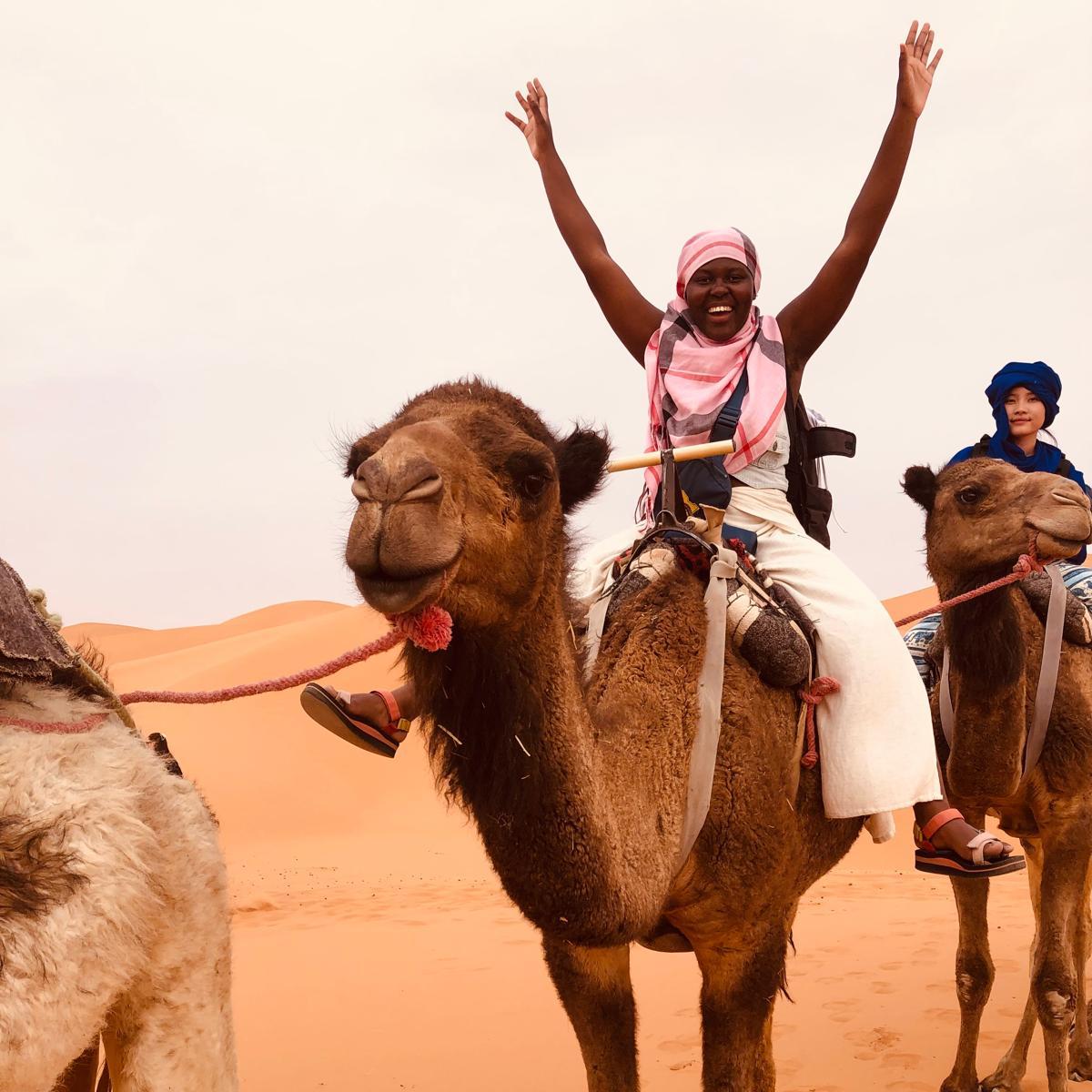A student riding on the back of a camel with her arms raised in the air in excitement