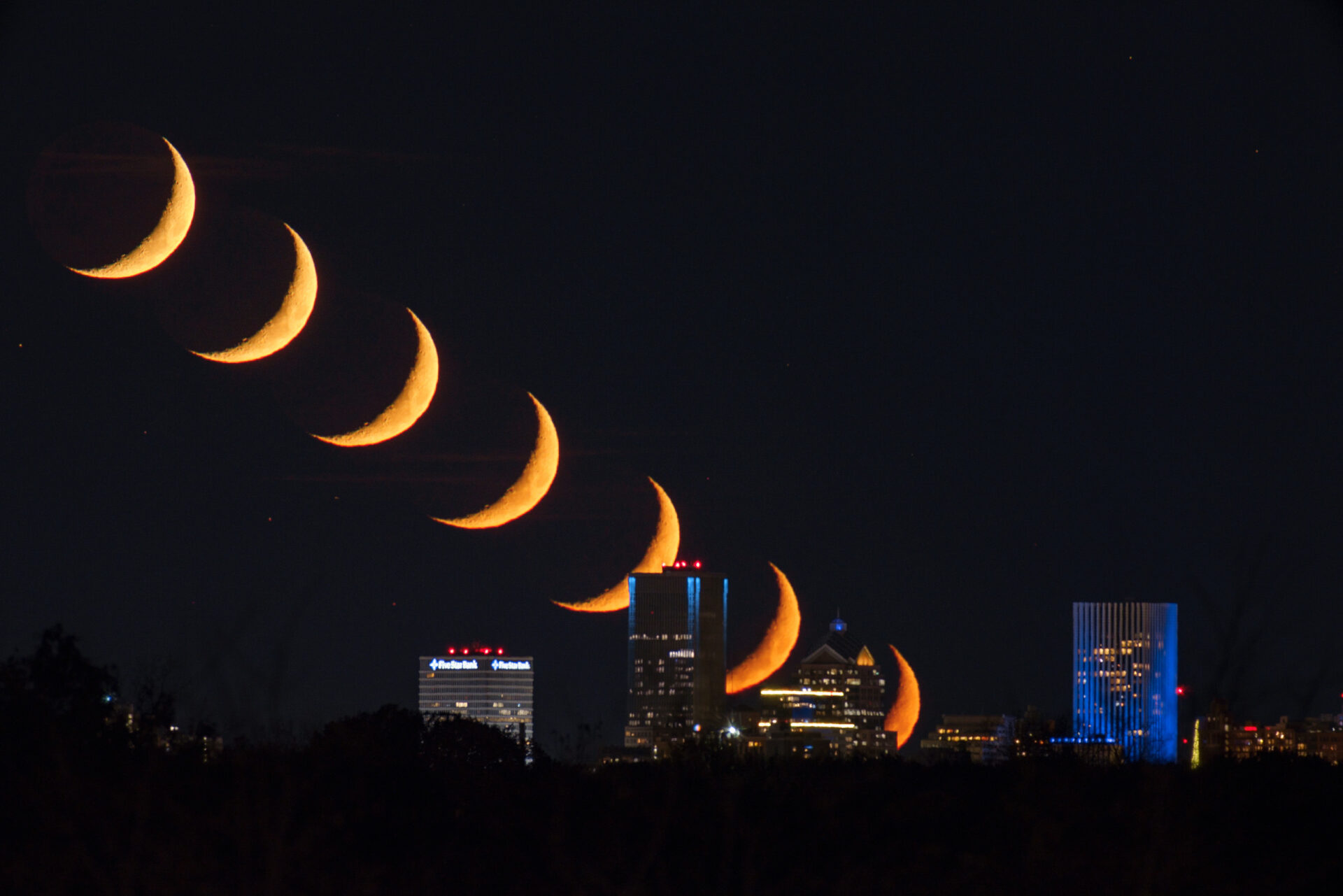 A time-lapse photo of the moon moving over the horizon line of downtown Rochester