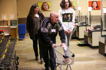 A man pushing a toy grocery shop and 2 women watching