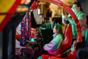 A female child plays a racing car arcade game as a woman takes a photo of her playing with her camera phone at the George Eastman Circle Family Celebration, held on April 27, 2023 within Rochester New York.