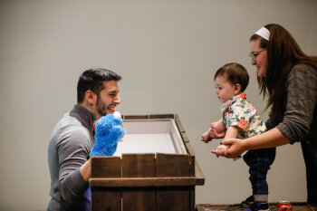 A candid photo of a man performing a puppet show for an infant whom is being held by a female at the George Eastman Circle Family Celebration, held on April 27, 2023 within Rochester New York.