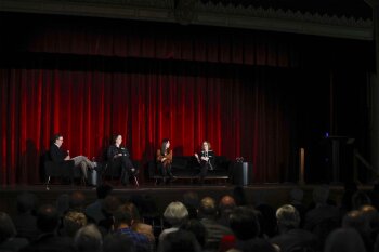 4 people seat on a stage in front of red curtains with audience in foreground