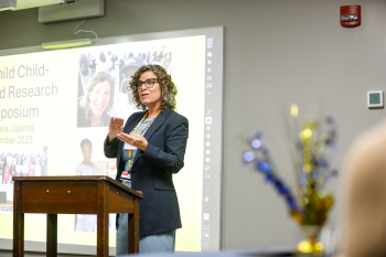 A woman at a lecturn speaking with a slide deck projecting in the background