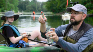 A man on kayak holding a pipette and jar with a woman in a kayak in the midground, on Genesee river.