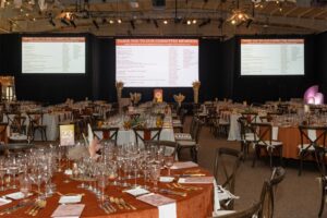 set tables with empty wine glasses and three projector screens with a list of the committee members