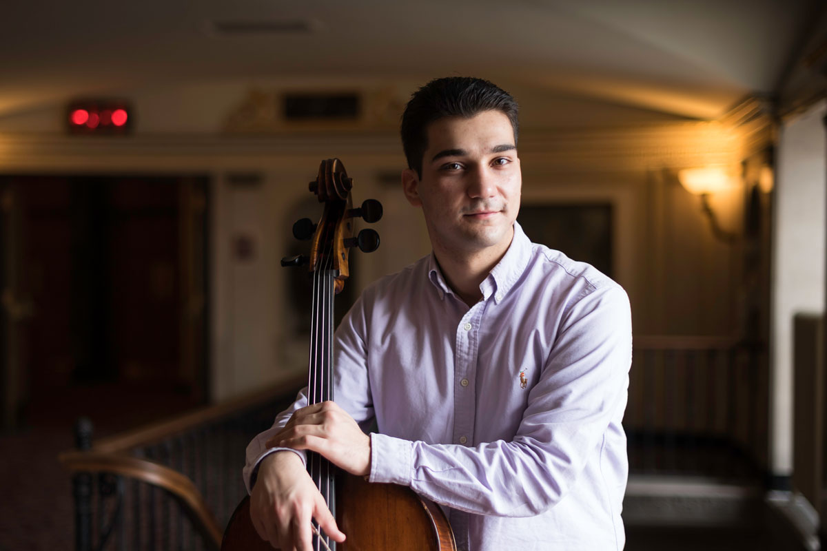 A University of Rochester student in a purple shirt holds a cello, showcasing his musical talent and dedication.