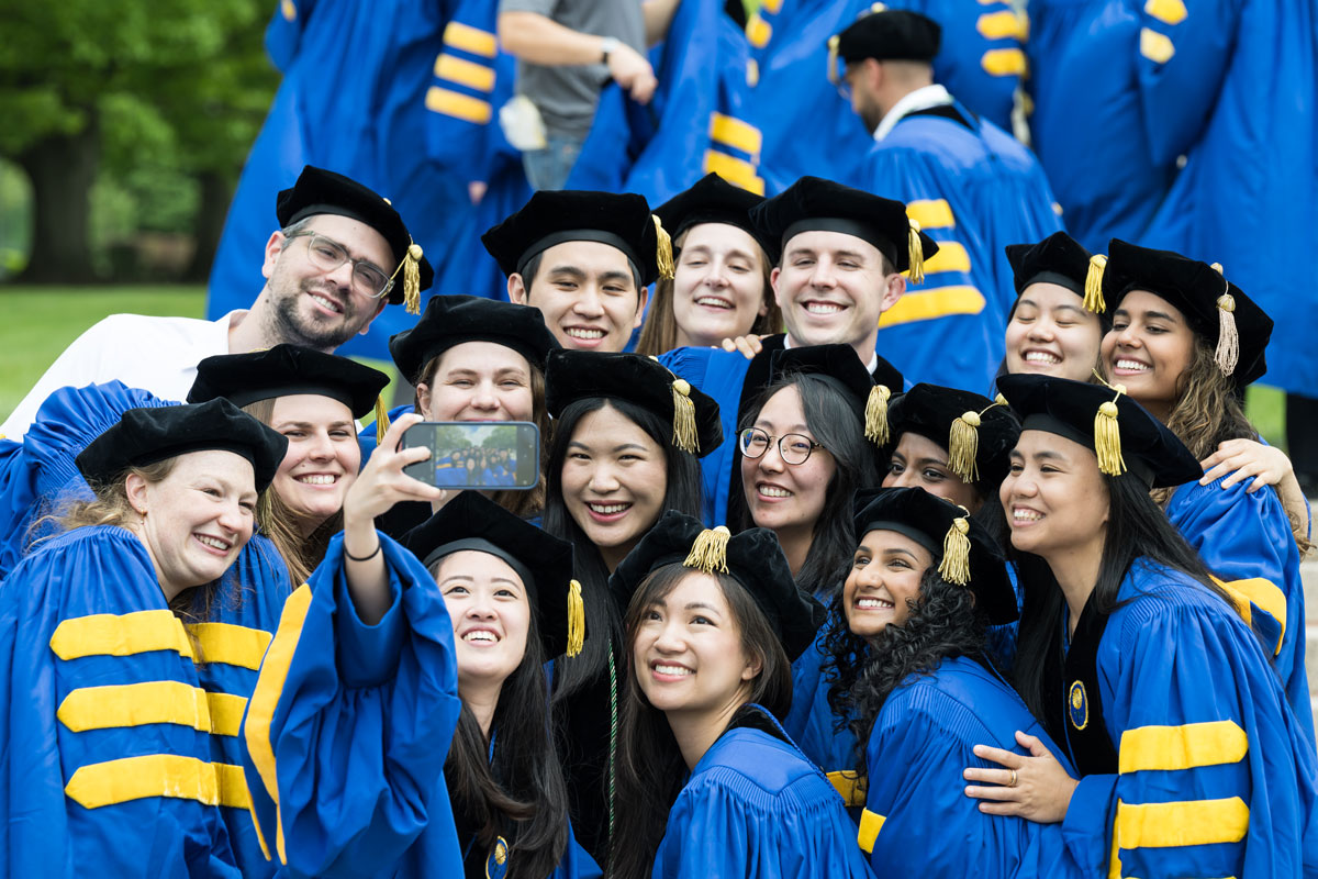 A group of University of Rochester graduates smiling and taking a selfie together in their caps and gowns.