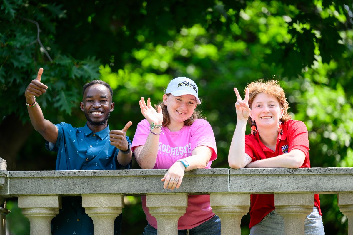 Three smiling students pose on a stone railing, making peace signs and thumbs-up gestures.