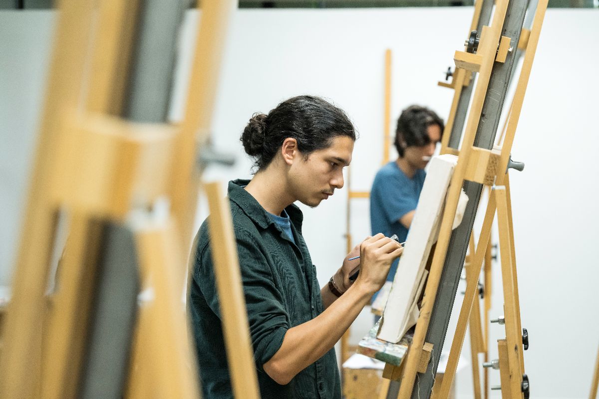 A University of Rochester student paints on a canvas in an art studio, surrounded by art supplies and natural light.