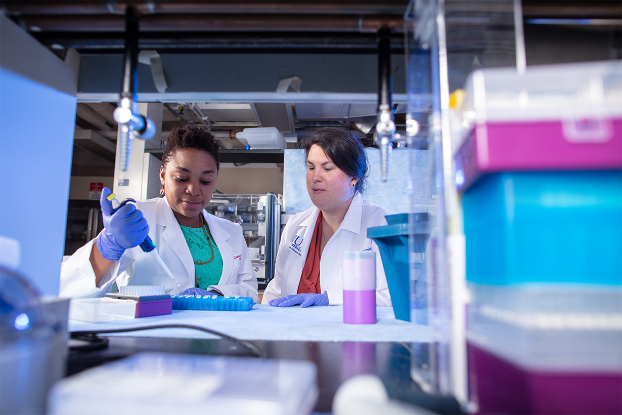 Two oncology researchers work together at a bench with a pipette.