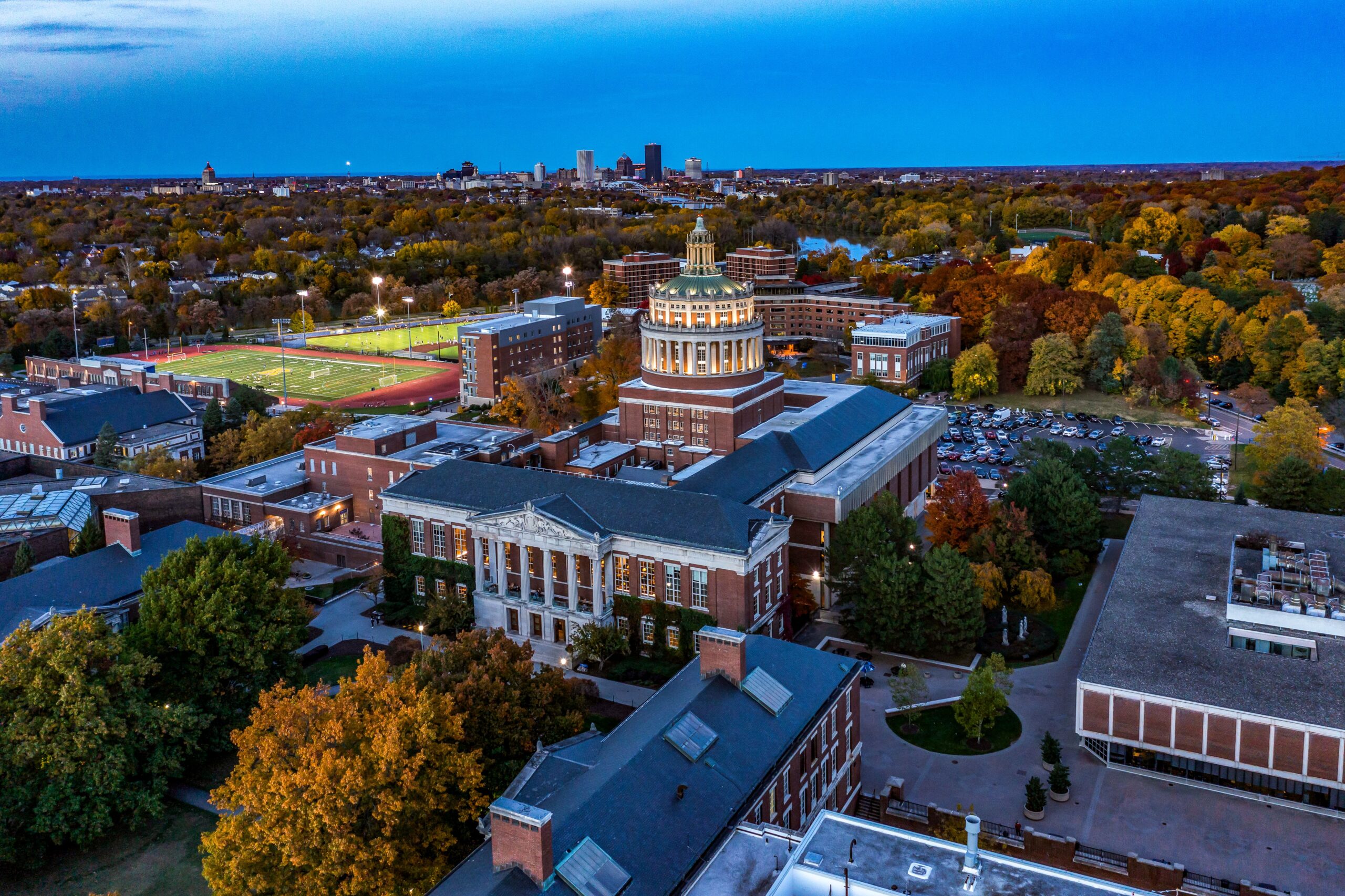 An aerial view of campus at dusk. Fall colors are seen on tress and the Rochester city skyline is visible in the upper left corner.