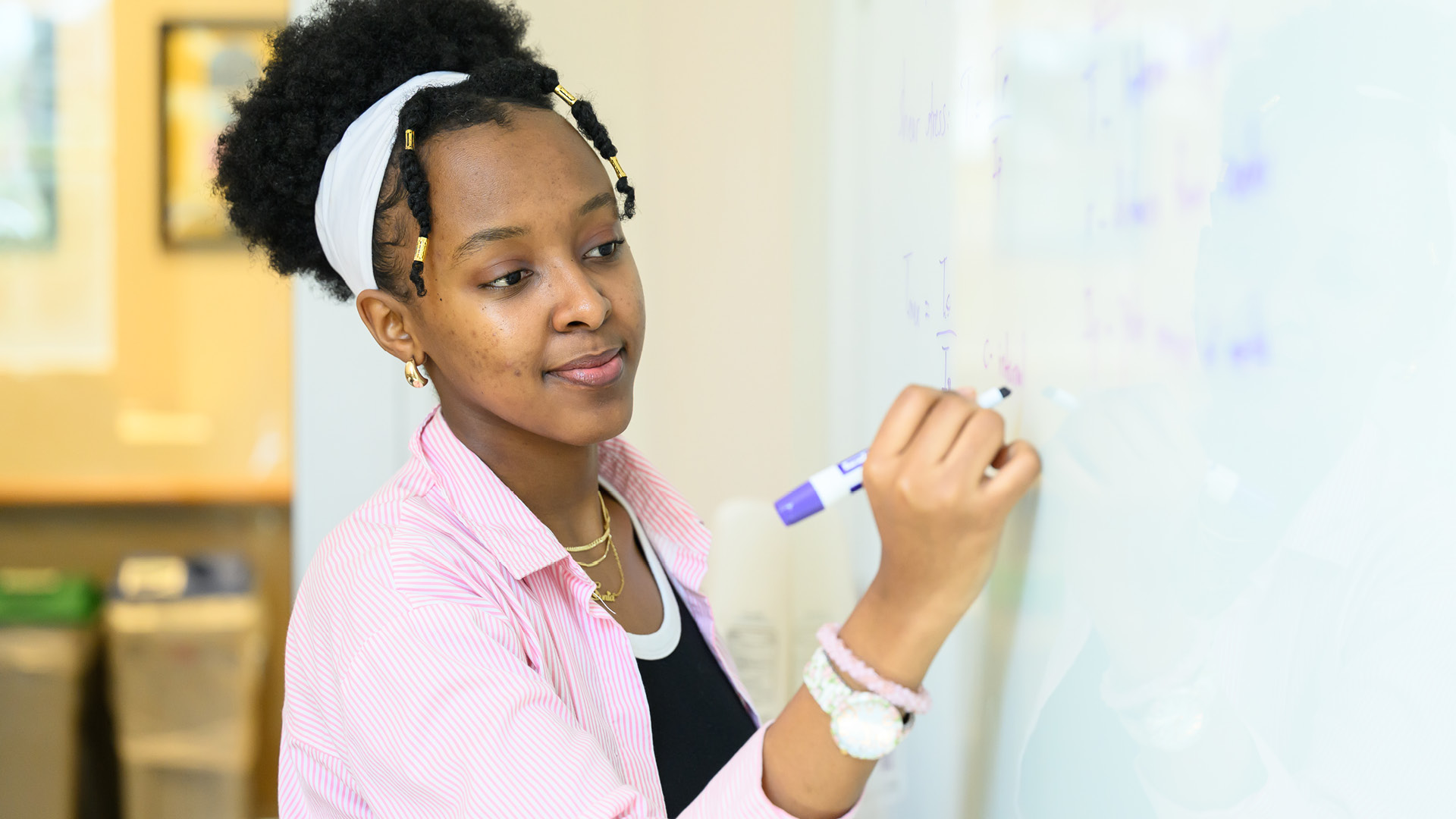 A student wearing a white headband and a pink blouse writes on a dry erase board with a blue marker.