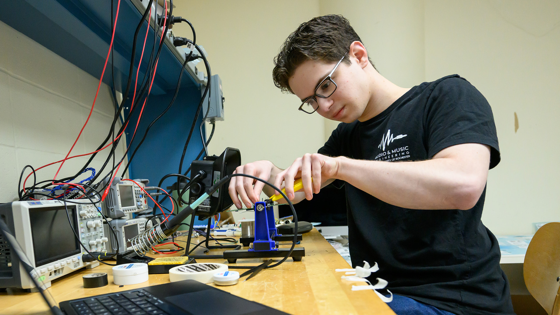 A male student in a black t-shirt and glasses works with wires in a lob setting.