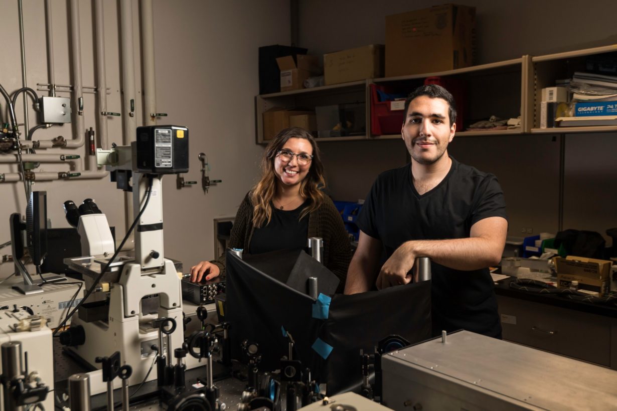 Two students in a research lab next to a giant microscope