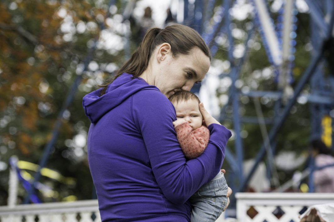 A young woman holding a baby in front of a ferris wheel at the University of Rochester's Meliora Weekend