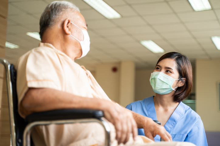 Asian senior psychiatric technician taking care of mature male patient sitting on wheelchair in hospital. Young woman and old man wearing surgical face mask for protection of The COVID pandemic. Girl talking to elderly man.