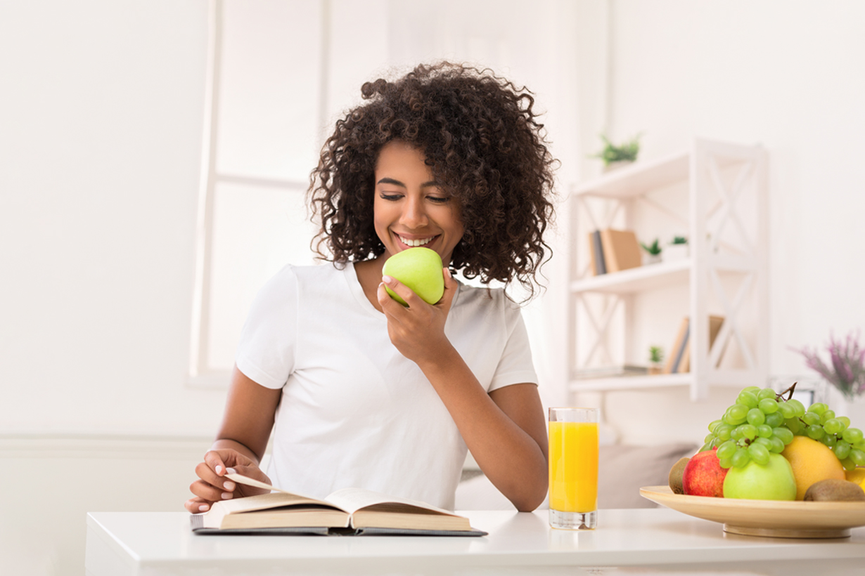 A woman sitting at a desk with one hand holding an apple while the other hand holds a page of a book.