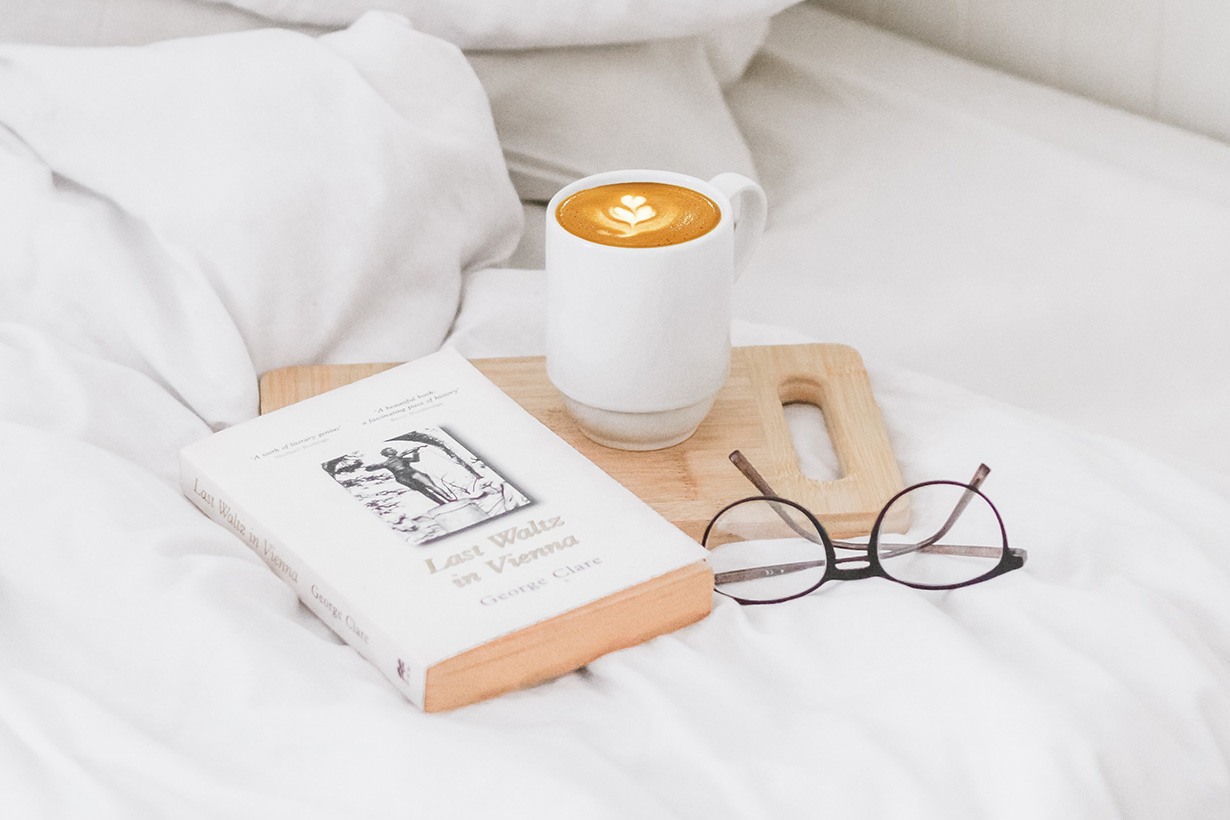 A cup, book, and pair of glasses on a bed.