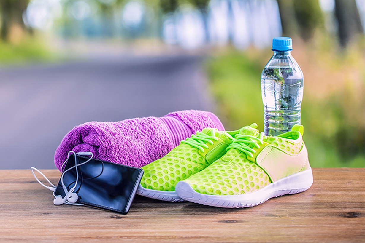 A pair of pink sneakers, water, a phone, and a purple towel are on a wooden bench.