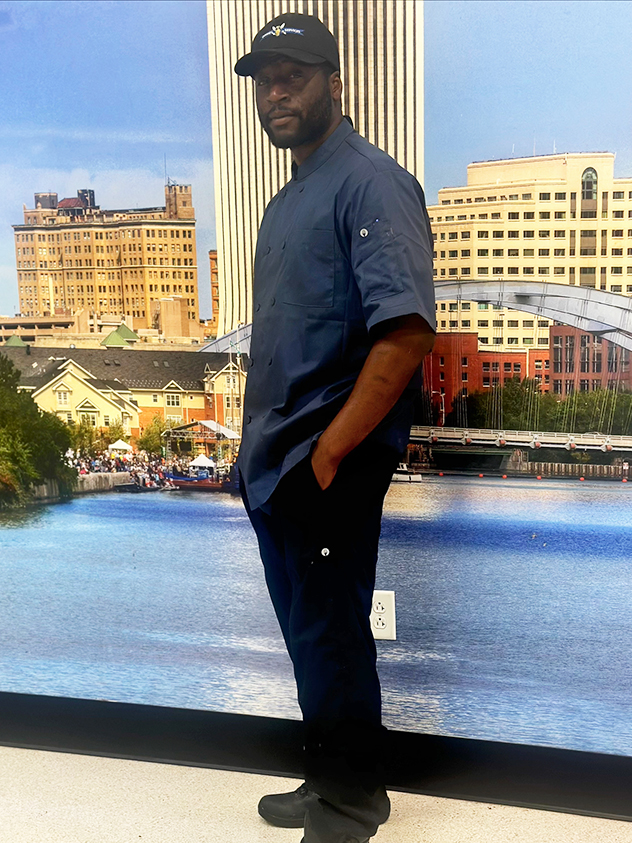 A man named JayQuan Coley, standing with one hand in his pant's pocket. He's wearing a baseball cap, dark blue button down and black pants.