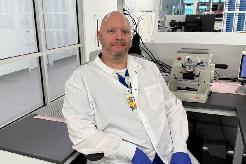 A man named Jason Toland, sitting on a chair with a white lab coat and blue gloves on. 