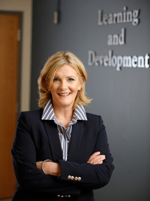 A woman named, Stepahnie VonBacho, standing in front of a wall with the text 'Learning and Development' in a blazer with her hands folded across her chest.