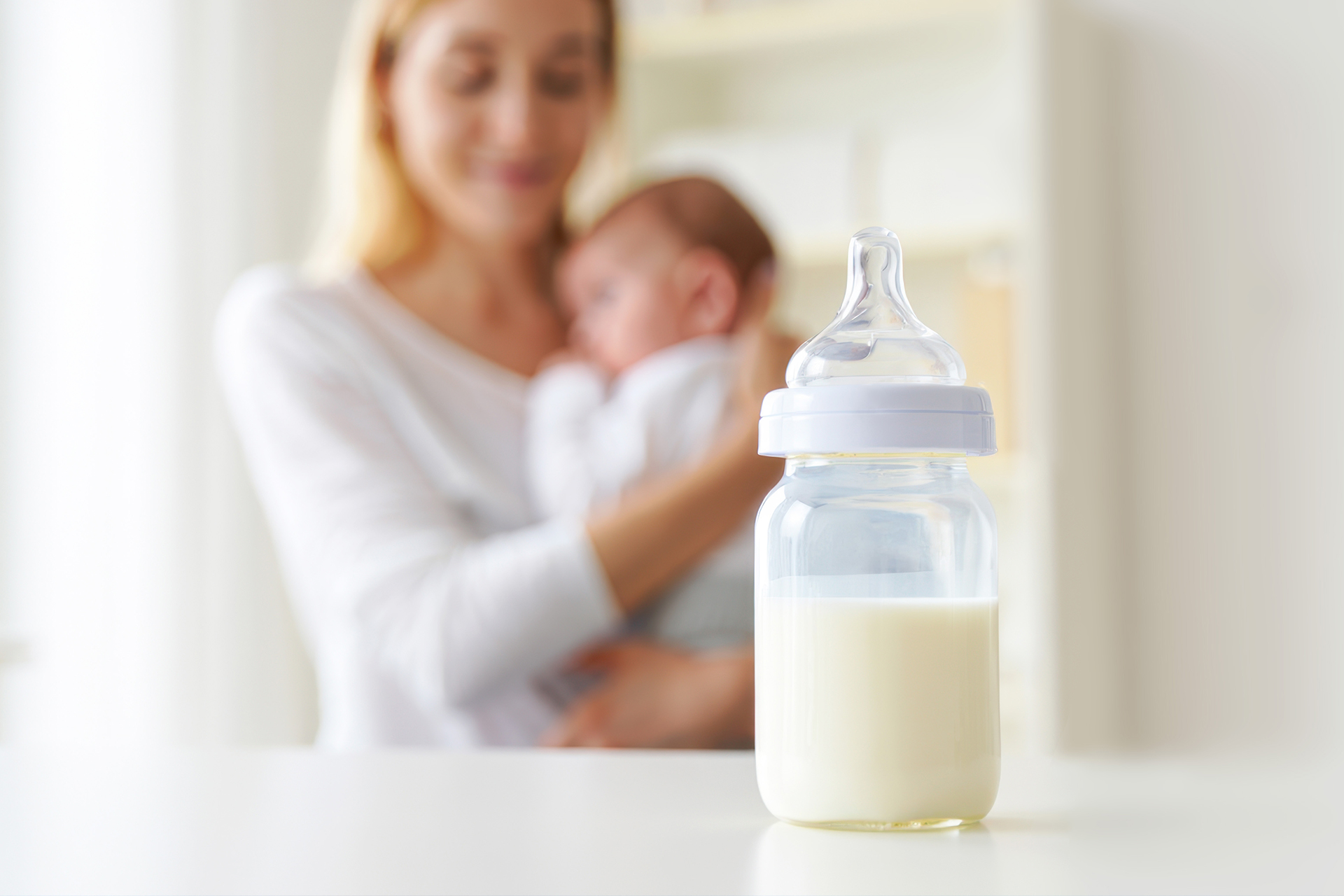 A baby bottle filled with milk in the foreground, with a caregiver holding an infant softly out of focus in the background.