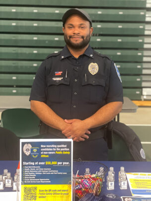 A man named Kamaran Moss, wearing a black Public Safety uniform, stands behind a table.