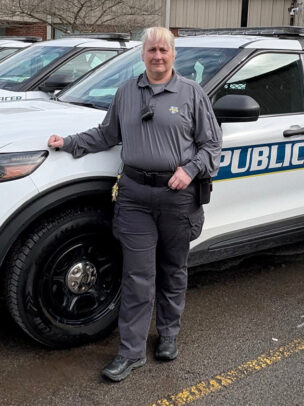 A woman named Sandra Rivers is standing against a public police van in a gray long-sleeve shirt and pants.