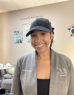 A woman named Leah Hill, wearing a hat, and smiling for the camera in her office space.