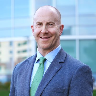 A man named Ryan Fields in a blue suit and green tie in front of a glass building.