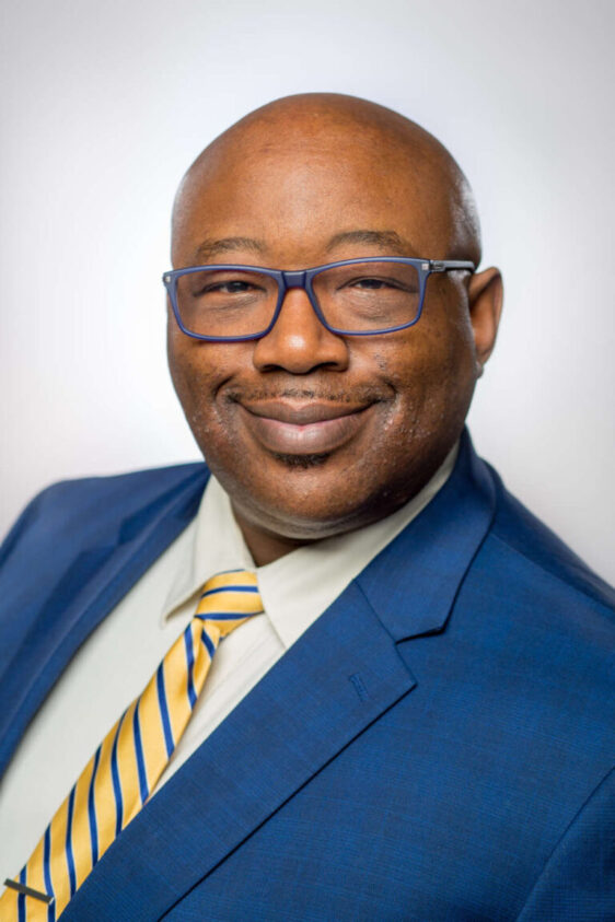 A man named John Blackshear smiles while wearing glasses, a blue blazer, and a yellow-and-blue striped tie.