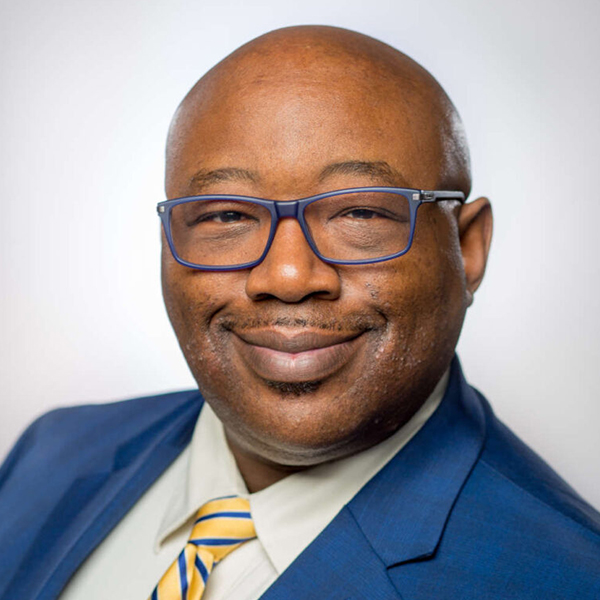 A man named John Blackshear smiles while wearing glasses, a blue blazer, and a yellow-and-blue striped tie.